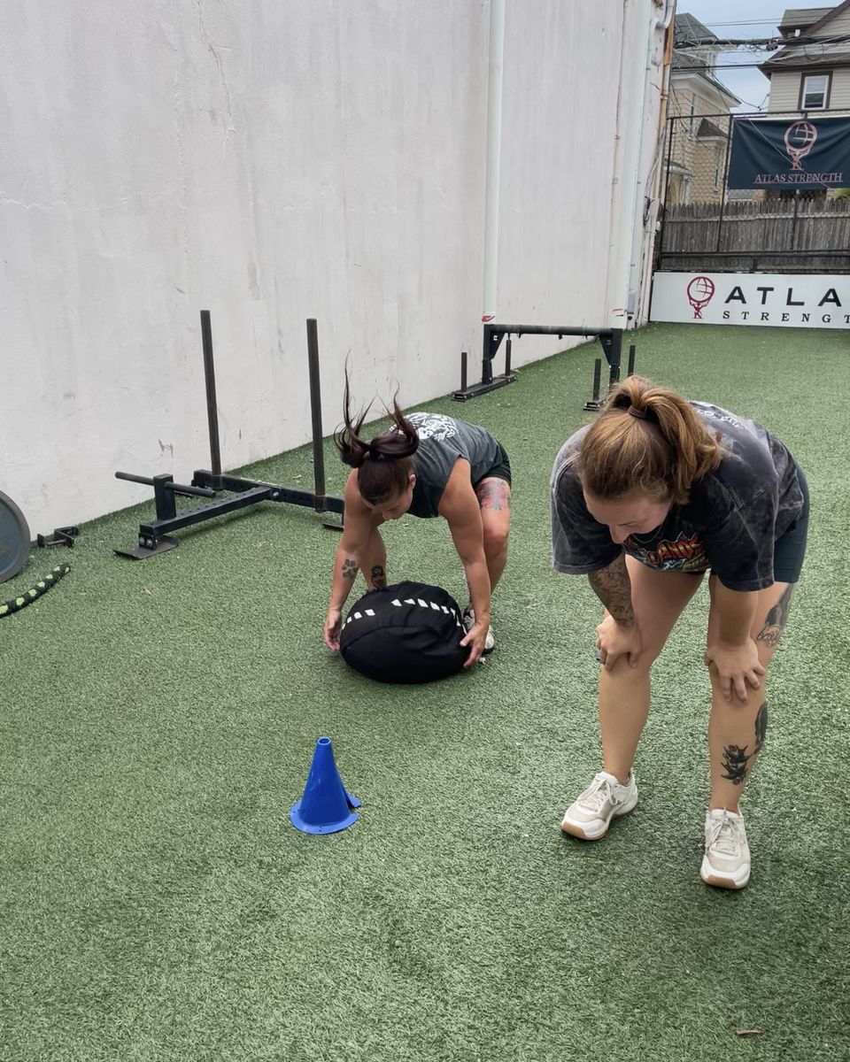 Two women training together with a sandbag on the outdoor turf