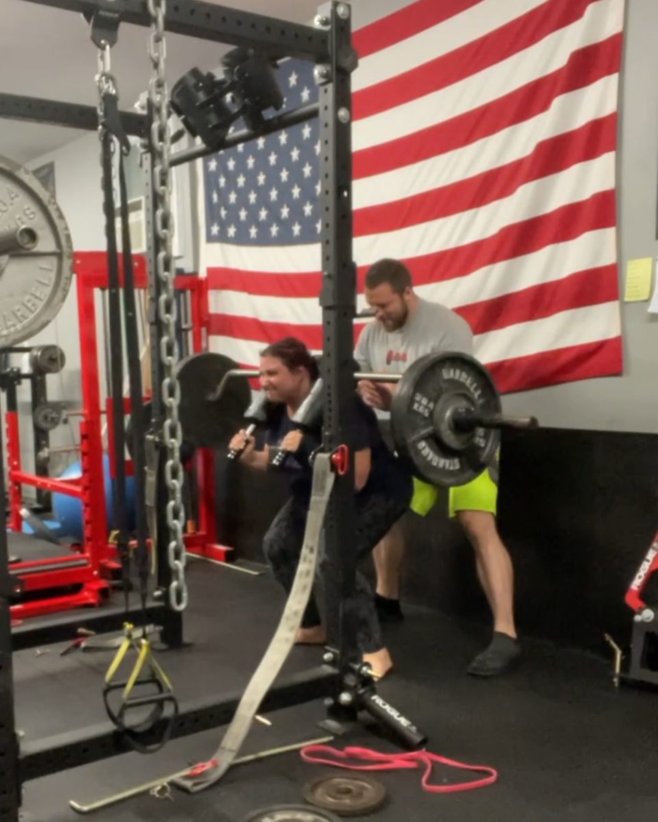 Woman squatting with a safety squat bar while Patrick Rice spots